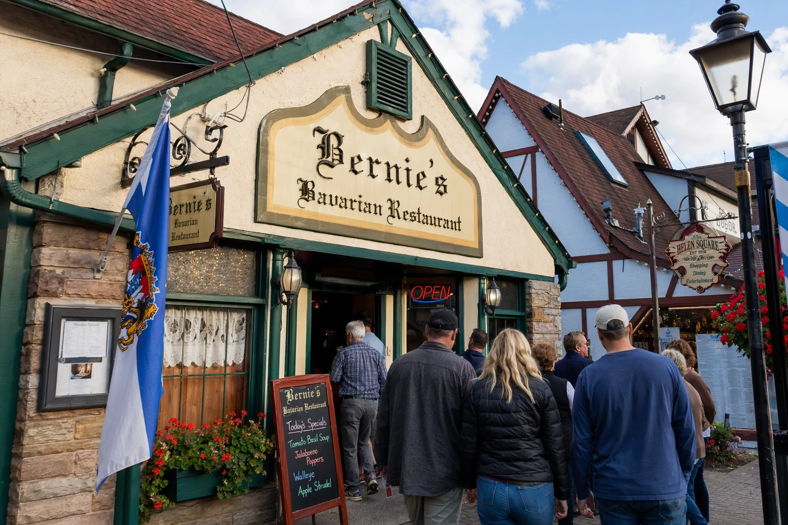 The exterior facade of Bernie's restaurant in Helen, Georgia, shot during the late afternoon when warm Georgia sunlight catches the painted signage above the entrance. The building sits along Helen's alpine-themed commercial district, where neighboring storefronts display Bavarian-style half-timbered trim and flower boxes. A sandwich board or menu stand near the front door lists daily specials, and the entrance is framed by the kind of low-key, locals-know-it signage that sets Bernie's apart from the flashier tourist spots along Bruckenstrasse. Foot traffic on the sidewalk suggests a busy lunch or early dinner hour, with visitors in casual outdoor clothing heading toward the door. The sky above is partly cloudy, giving the scene even, diffuse light that brings out the color of the storefront paint and surrounding alpine architectural details.