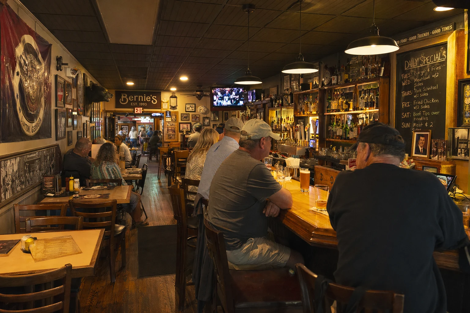 Inside Bernie's in Helen, Georgia, the dining room shows a casual, no-frills setup with wooden tables and chairs arranged along a narrow space. The walls carry memorabilia, handwritten daily specials on a chalkboard, and perhaps a few pieces of local Georgia mountain decor. Pendant lights or recessed fixtures cast warm yellow light over the bar counter and dining tables, giving the room an intimate tavern feel that contrasts with the touristy alpine streetscape just outside the windows. A few patrons sit at the bar with glasses of beer or cocktails in front of them. Behind the bar, shelves hold bottles organized in rough rows, and a tap handle or two is visible at counter height. The overall impression is a relaxed neighborhood hangout embedded inside a town built largely for visitors.