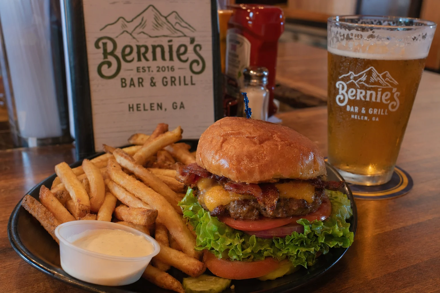 A plate of food served at Bernie's in Helen, Georgia, photographed on a wooden or laminate tabletop in the restaurant's interior lighting. The dish reflects the casual American comfort-food menu typical of a local Helen bar and grill, possibly a burger stacked with lettuce, tomato, and cheese on a toasted bun, or a basket of seasoned fries with a dipping sauce on the side. The portion size is generous, consistent with a spot that caters to hungry hikers and tubers coming off the Chattahoochee River nearby. The plate sits next to a cold pint glass of draft beer, condensation visible on the outside of the glass. The image communicates honest, straightforward food at a Helen local institution rather than fine dining presentation.