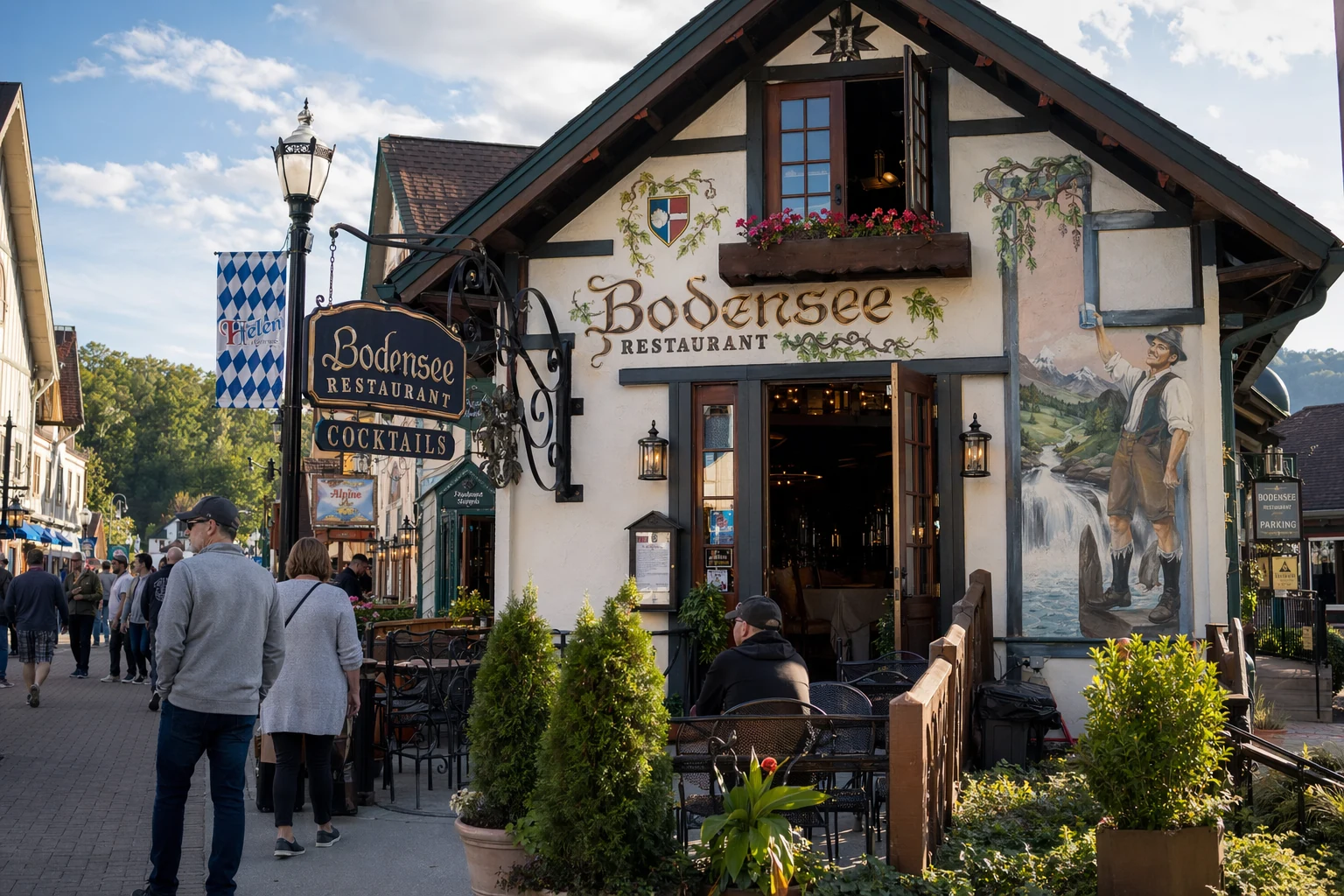 Bodensee restaurant exterior in Helen, Georgia, displaying distinctive Bavarian alpine architecture with decorative white stucco walls and dark timber framing. The building features traditional Germanic design elements including a painted facade and flower boxes beneath windows. Located along Helen's alpine village streetscape, the restaurant sits among other themed buildings that recreate a southern German mountain town atmosphere. Afternoon sunlight illuminates the front entrance and outdoor seating area. Visible signage identifies the establishment, while surrounding businesses and pedestrian walkways create the immersive European village setting that defines Helen's tourism district in the north Georgia mountains.