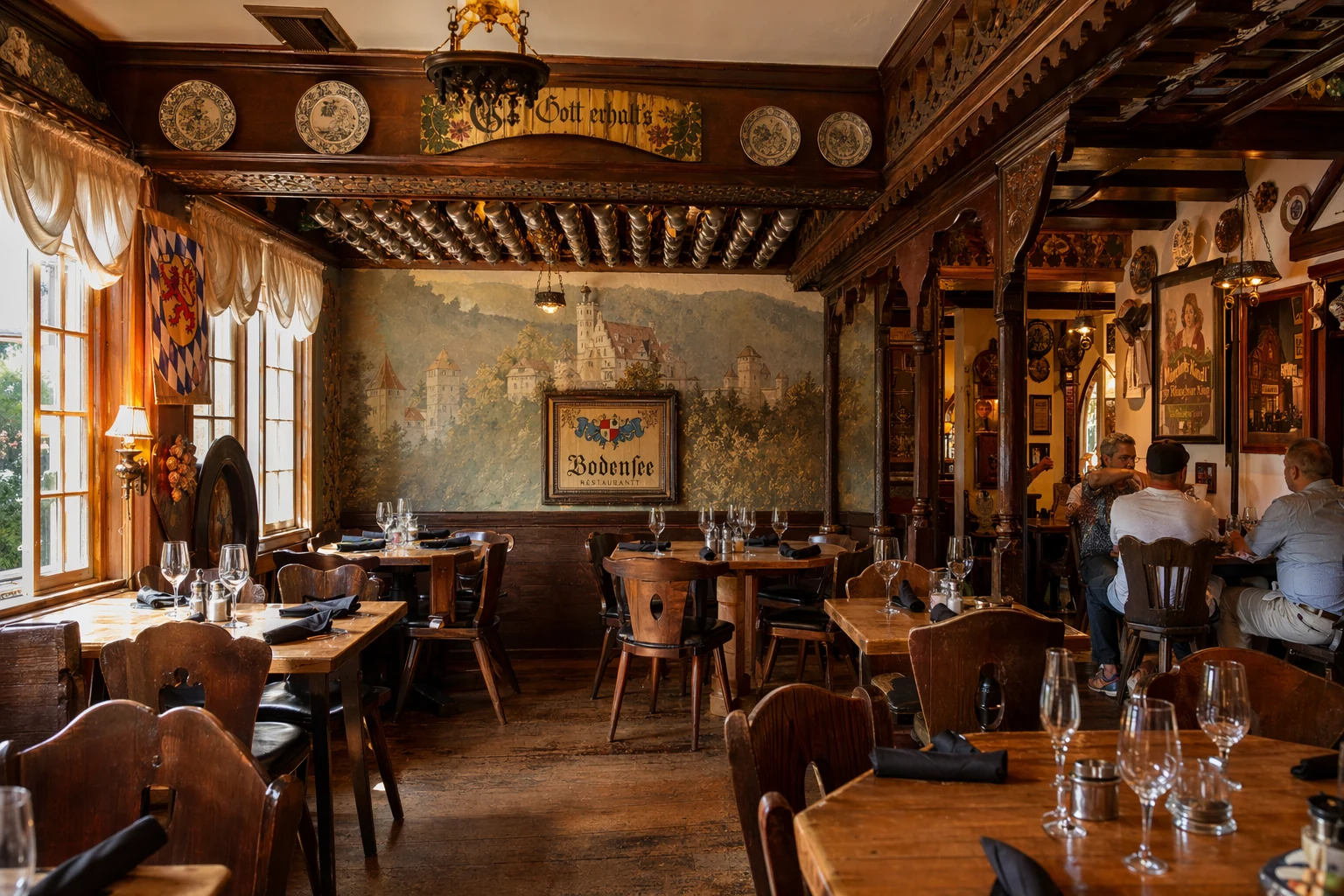 Interior dining room at Bodensee in Helen, Georgia, showing wooden tables and chairs arranged throughout the space with Bavarian themed decor on walls and ceilings. The room features dark wood paneling, traditional German accents, and warm ambient lighting that creates an authentic biergarten atmosphere. Dining tables are set with glassware and napkins, ready for guests. Windows allow natural light to filter into the space during daytime service. The interior design continues the alpine village theme found throughout Helen, with attention to Germanic cultural details that transport diners to a European mountain lodge setting in the heart of the north Georgia foothills.