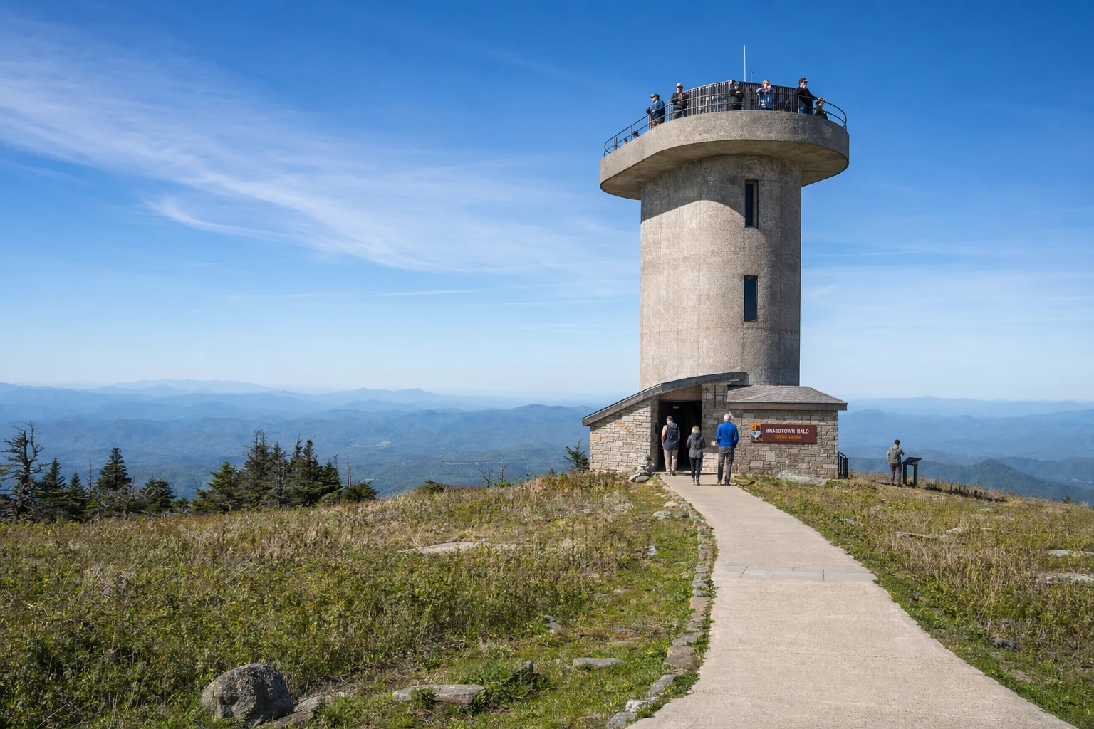 Brasstown Bald observation tower stands at the summit of Georgia's highest peak near Helen. The concrete tower rises above the mountain's rounded bald top, with visitors visible on the observation deck scanning the horizon. A paved walkway leads to the tower entrance, surrounded by low vegetation and exposed rock typical of this alpine environment. The sky stretches wide and blue overhead, while multiple mountain ridges fade into the distance in layers of blue and green. At 4,784 feet elevation, this tower provides 360-degree views across four states. The National Forest Service maintains this site as a key destination along the Appalachian highlands. Late morning sunlight illuminates the structure and the open meadow surrounding it.