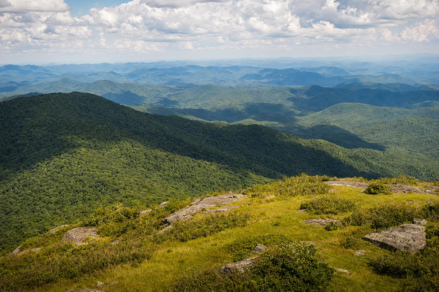 The four-state view from Brasstown Bald summit stretches across endless Appalachian ridges near Helen, Georgia. Layer upon layer of mountain ranges recede into the horizon, each ridge appearing progressively lighter in shades of blue and gray due to atmospheric perspective. The foreground shows the grassy bald mountaintop with scattered low shrubs and exposed granite. Dense hardwood forest covers the slopes below, showing the rich green of summer growth. On clear days, visitors can see into North Carolina, Tennessee, and South Carolina from this vantage point. Puffy white clouds cast moving shadows across the distant valleys. The vast scale of the southern Appalachian Mountains becomes evident from this highest point in Georgia, where the landscape unfolds in all directions.