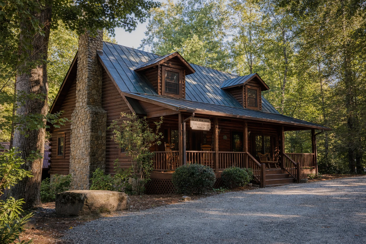 A wooden log cabin rental sits nestled among tall hardwood and pine trees in Helen, Georgia. The structure features a traditional stacked log construction with dark stain, a metal roof, and a wide front porch with wooden rocking chairs. A stone chimney rises along one side of the building. Dappled sunlight filters through the forest canopy, casting shadows across the cabin's facade. The surrounding landscape shows native mountain vegetation and a gravel driveway leading to the entrance. This vacation rental exemplifies the rustic Appalachian architecture common to the North Georgia mountains, offering visitors a secluded retreat within minutes of Helen's alpine village center.