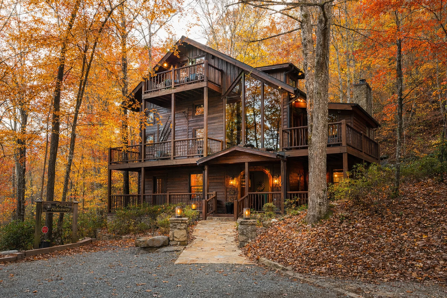 A cabin rental in Helen, Georgia appears through autumn foliage, with surrounding trees displaying vibrant orange, red, and yellow leaves. The wood sided cabin features multiple levels built into the hillside, with expansive windows reflecting the fall colors. A stone pathway leads from a parking area to the main entrance. The forest floor is carpeted with fallen leaves. This seasonal view demonstrates the appeal of Helen area cabin rentals during fall, when the hardwood forests of the Chattahoochee National Forest transform into brilliant color displays, drawing visitors to the North Georgia mountains for leaf peeping and cooler mountain temperatures.