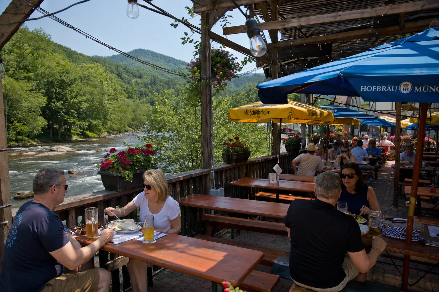 An outdoor biergarten in Helen, Georgia on a sunny afternoon, with long wooden picnic tables and benches arranged under a pergola structure. Colorful market umbrellas in blue and yellow provide shade over some tables. String lights crisscross overhead between wooden posts. Guests sit with beer mugs and plates of food, surrounded by potted geraniums and hanging flower baskets. The Chattahoochee River flows visibly in the background beyond a low wooden fence. Forested mountains rise green in the distance under a clear sky. This relaxed outdoor dining setting allows visitors to enjoy German beer and food while taking in the natural beauty of the North Georgia mountains.