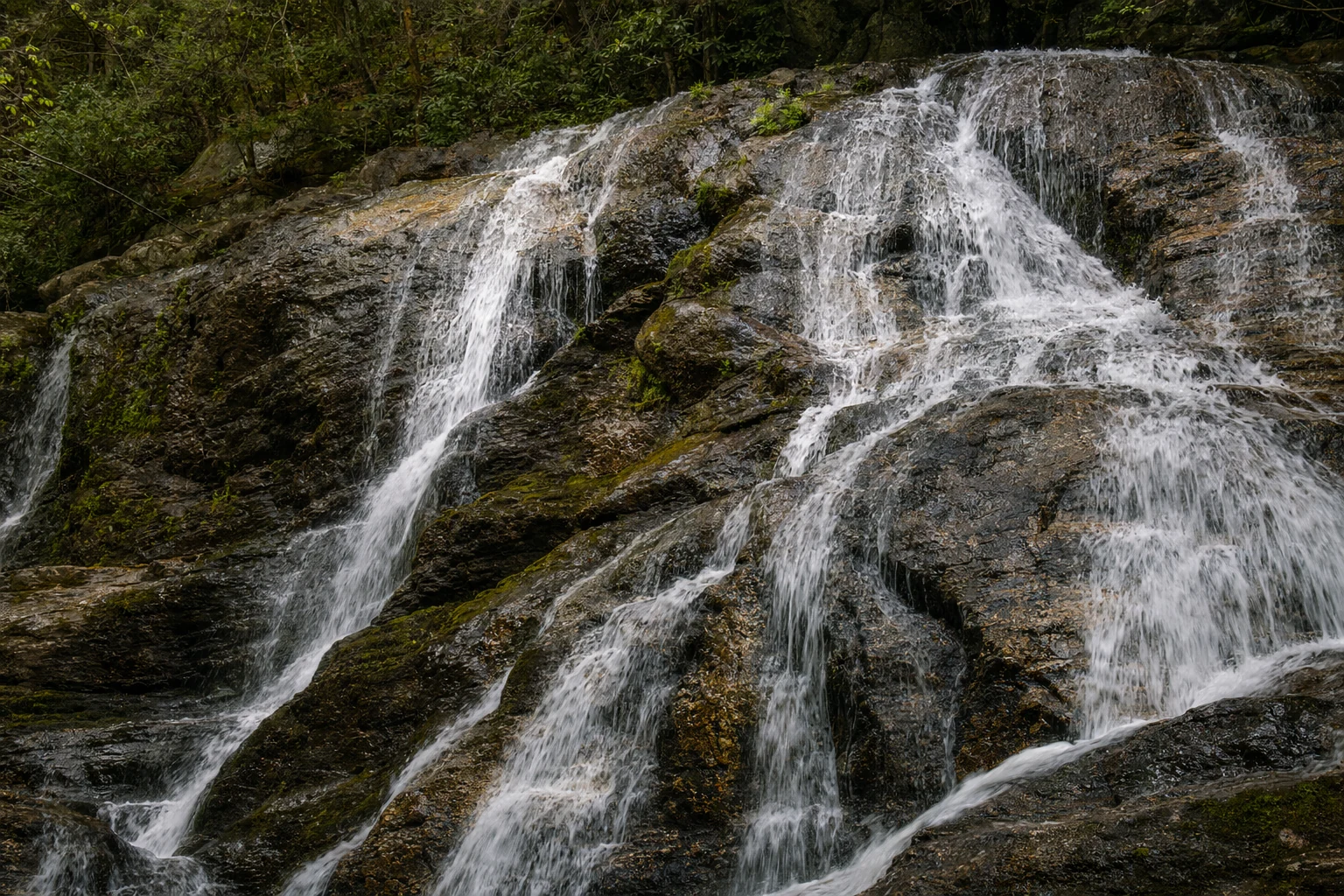Close view of water flowing over the rocky cascade at Dukes Creek Falls in the Helen, Georgia area, showing the texture and movement of the multi-tiered waterfall. White water rushes over dark gray and rust-stained granite ledges, creating sheets and ribbons of flow that catch available light. Smaller cascades branch and rejoin as gravity pulls the creek downward through narrow chutes and over rounded stone edges worn smooth by centuries of water movement. Green moss and lichen cling to rocks at the waterfall edges where constant spray keeps surfaces damp. The surrounding forest remains slightly out of focus in the background, emphasizing the dynamic water movement and geological features of this popular north Georgia waterfall.
