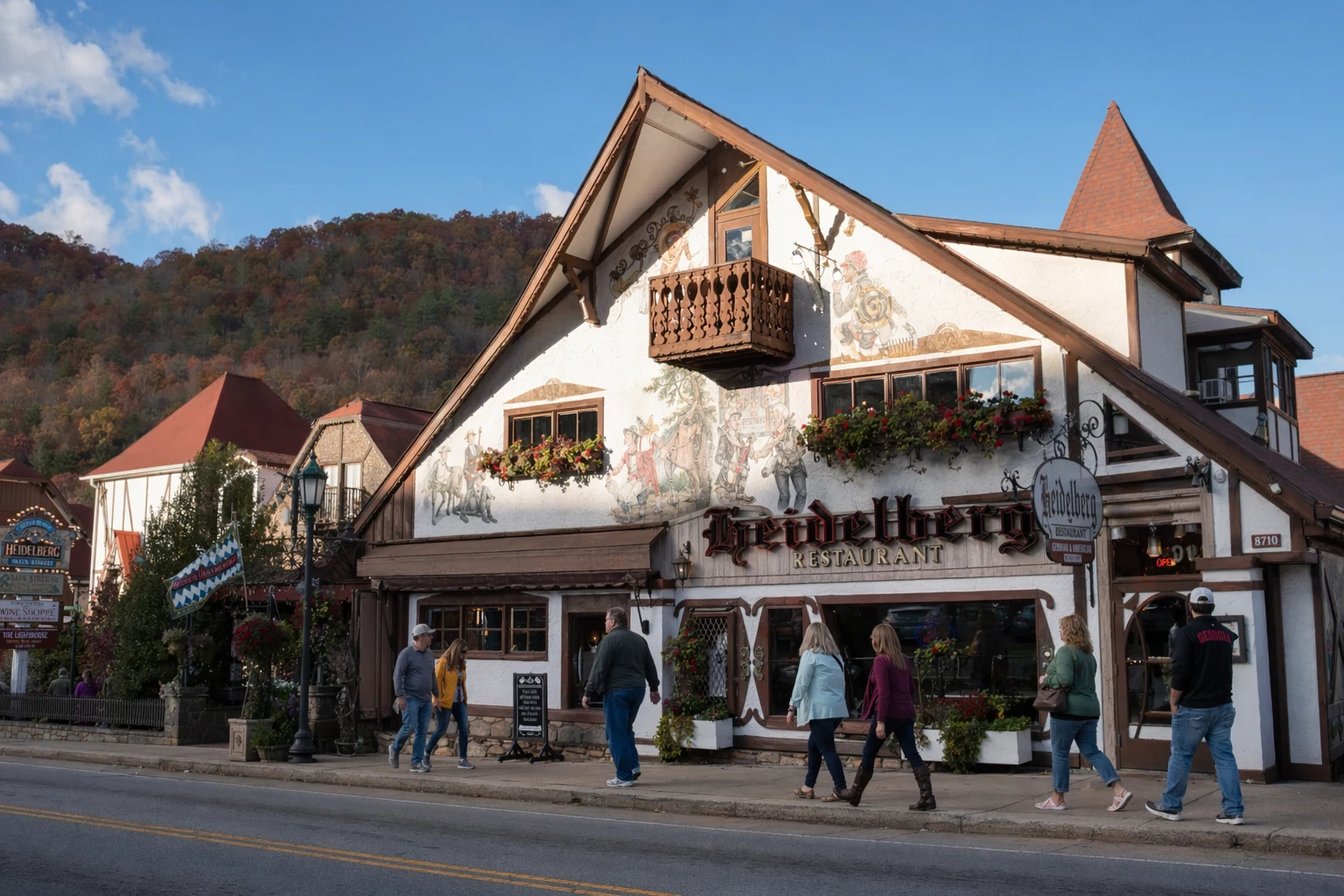 Heidelberg restaurant stands along the main street in Helen, Georgia, featuring traditional Bavarian alpine architecture with a steeply pitched roof, decorative wooden balconies, and colorful murals painted across the facade. The building displays ornate German folk art and bright flower boxes beneath second-story windows. Visitors walk along the sidewalk in front of the restaurant, which sits among other alpine-style buildings that define Helen's reimagined German village atmosphere. The exterior showcases rich earth tones of brown timber framing against cream stucco walls, with signage in Gothic lettering identifying the establishment. Blue sky and mountain ridges form the backdrop to this recreation of a European village streetscape in the north Georgia mountains.