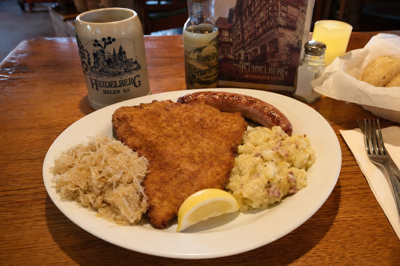 A traditional German platter served at Heidelberg restaurant in Helen, Georgia presents schnitzel, sausages, sauerkraut, and potato salad arranged on a white oval plate. The golden breaded schnitzel cutlet occupies the center, accompanied by grilled bratwurst or knockwurst with browned casing. Tangy sauerkraut and creamy German potato salad fill the remaining plate space. A lemon wedge garnishes the schnitzel. The meal sits on a wooden table inside the restaurant, representing authentic Bavarian cuisine adapted for American visitors to this alpine-themed north Georgia town. The portion size is generous, reflecting the hearty mountain dining tradition. This combination plate showcases the central European culinary heritage that Heidelberg brings to the Helen dining scene.