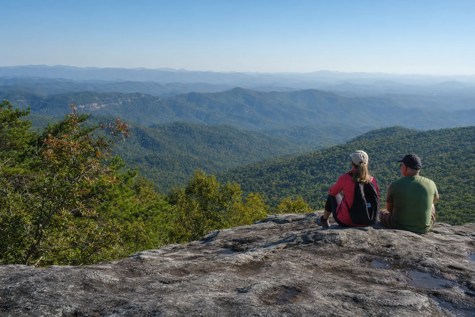 A sweeping mountain vista from an overlook near Helen, Georgia shows layers of blue ridges receding into the distance under a clear sky. The foreground features a rocky outcrop where hikers can rest and take in the panoramic view of the Chattahoochee National Forest spreading across the valleys below. Multiple mountain ranges create a classic Blue Ridge effect with each successive ridge appearing lighter and hazier in the atmospheric perspective. Dense forest cover blankets the slopes in various shades of green, with occasional clearings or rock faces breaking the tree line. This viewpoint represents the reward found at the end of several challenging trails accessible from Helen, where elevation gain provides visitors with spectacular views of the Southern Appalachian landscape stretching across Northeast Georgia.